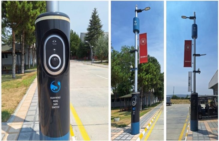 Görselde, bir sokak üzerinde konumlandırılmış modern teknolojiyle donatılmış aydınlatma direkleri görülüyor. Direklerin alt kısmında “Elektrikli Araç Şarj Ünitesi” yer almakta ve Togg logosu dikkat çekiyor. Direklerin üst bölümlerinde ise sokak lambaları ve çeşitli sensörler bulunuyor. Ayrıca direklere Enerji ve Tabii Kaynaklar Bakanlığı'nı temsil eden kırmızı bayraklar asılmış.