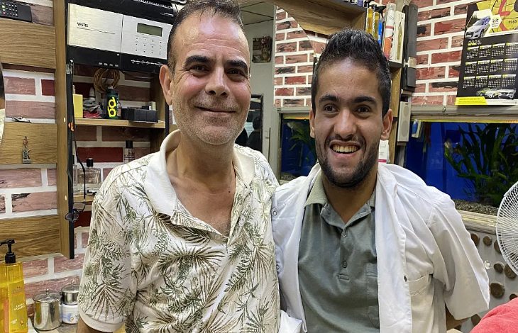 Görselde, bir berber dükkanında Ayhan Deliorman ve zihinsel engelli birey Faruk Taymura yan yana ayakta duruyor.