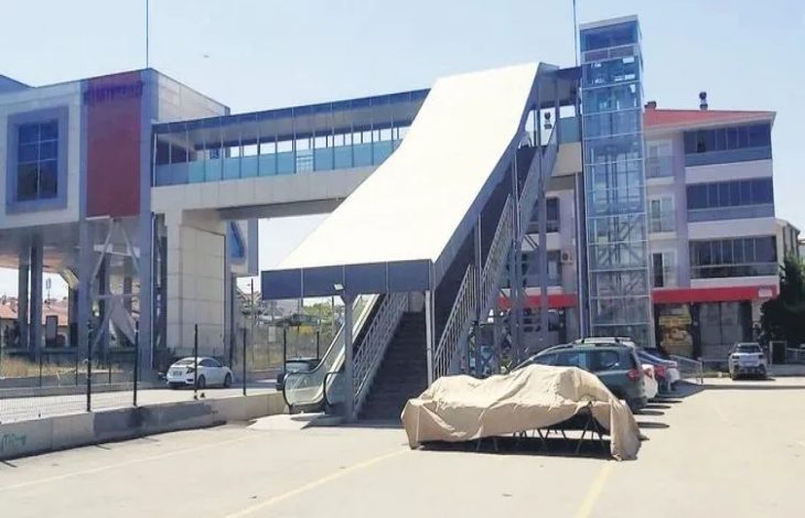 Görselde, bir üstgeçit ve ona bağlı asansör ile yürüyen merdiven sistemi görülüyor. Ancak merdivenlerin çalışmadığı ve üstünün tente ile kapatıldığı anlaşılıyor. Sol tarafta yürüyen merdivenler, sağda ise cam yapılı bir asansör var. Alt kısımda birkaç araç park halinde ve üst geçidin bulunduğu bina modern yapıda.