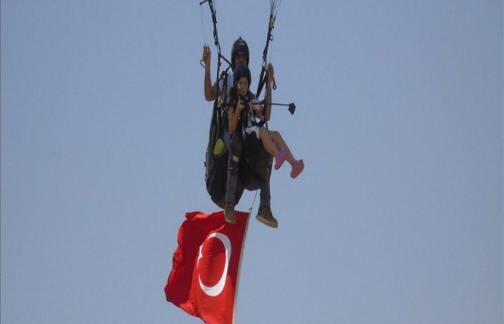 Fotoğrafta, gökyüzünde yamaç paraşütü yapan iki kişi görülüyor. Önde oturan genç kadın, arkasında ise paraşüt pilotu yer alıyor. İkilinin altında büyük bir Türk bayrağı dalgalanıyor. Arka plan tamamen mavi gökyüzünden oluşuyor.
