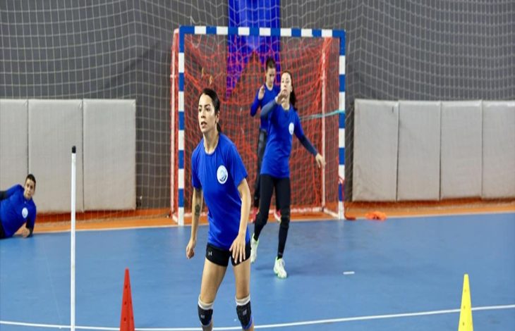 Görselde, mavi tişört ve dizlik takmış bir kadın hentbolcu Kübra Akçayoğlu salon içinde antrenman yapıyor. Ön planda sporcu koşarken, arka planda kalenin önünde ve yanında duran diğer takım arkadaşları görülüyor. Zeminde antrenman için yerleştirilmiş turuncu ve sarı koniler yer alıyor.