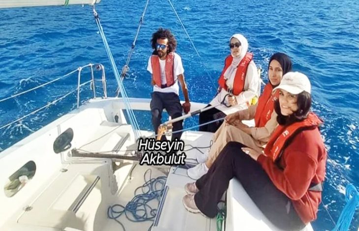 Görselde, deniz üzerinde seyreden bir yelkenlide dört kişi yer alıyor. Hepsi can yeleği giymiş ve teknenin içinde oturuyor. Ön planda üç kadın, arka tarafta ise dümeni tutan ve adı görselde “Hüseyin Akbulut” olarak belirtilen bir erkek bulunuyor. Masmavi deniz ve açık hava, keyifli bir yelken yolculuğu atmosferi oluşturuyor.