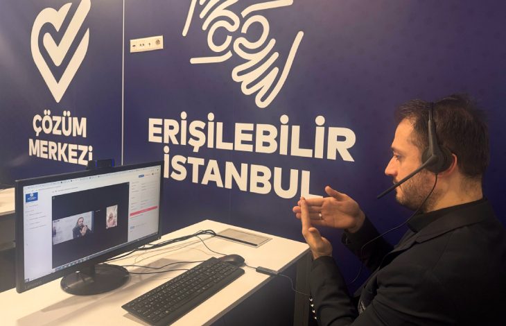 Fotoğrafta, bir erkek bilgisayar başında oturuyor ve kulaklık-mikrofon seti takılı. Ekranda çevrim içi bir görüşme gerçekleştiriliyor. Erkek kişi, ellerini kullanarak işaret diliyle iletişim kuruyor. Duvarın arkasında beyaz renkte büyük harflerle "ERİŞİLEBİLİR İSTANBUL" ve "ÇÖZÜM MERKEZİ" yazıları yer alıyor. Aynı zamanda işaret diliyle "erişilebilir" kelimesini temsil eden bir simge de duvarda bulunuyor. Masa üzerinde bir bilgisayar monitörü, klavye, fare ve cep telefonu yer alıyor. Görsel, İstanbul'da engelliler için erişilebilirlik ve iletişim destek hizmetleri sağlandığını gösteren bir ortamı yansıtıyor.