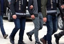 Görselde, polis yeleği giymiş ekipler kolundan tuttukları kişiyi götürüyor. Yan yana yürüyen birkaç polis ve sivil kişi görülüyor, ortam açık hava ve araçlar arka planda yer alıyor.