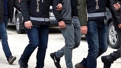 Görselde, polis yeleği giymiş ekipler kolundan tuttukları kişiyi götürüyor. Yan yana yürüyen birkaç polis ve sivil kişi görülüyor, ortam açık hava ve araçlar arka planda yer alıyor.