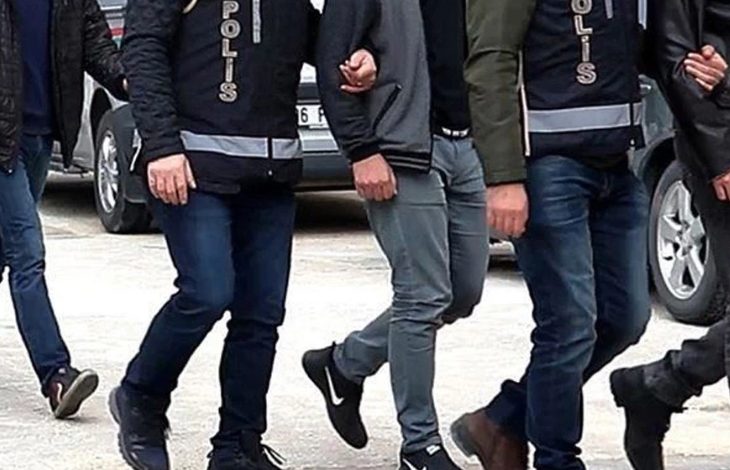 Görselde, polis yeleği giymiş ekipler kolundan tuttukları kişiyi götürüyor. Yan yana yürüyen birkaç polis ve sivil kişi görülüyor, ortam açık hava ve araçlar arka planda yer alıyor.