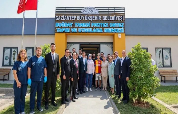 Görselde, Gaziantep Büyükşehir Belediyesi’ne ait “Buğday Tanesi Protez Ortez Yapım ve Uygulama Merkezi” önünde toplanmış kalabalık bir grup yer alıyor. Kadın ve erkeklerden oluşan ekip, resmi kıyafetli yetkililer, sağlık personelleri ve sivil vatandaşlardan oluşuyor. Ortada, Galatasaray forması giyen bir çocuk ve ailesi bulunuyor; büyük olasılıkla protez kol uygulaması yapılan çocuk bu. Arka planda merkezin adı tabelada mavi harflerle yazılı.