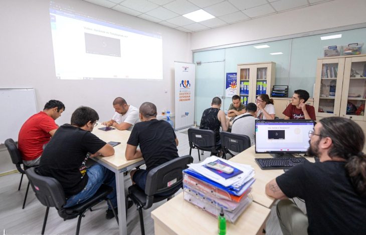 Katılımcılar, iki ayrı masa etrafında oturmuş durumda ve çoğunlukla tablet veya defter gibi materyallerle meşguller. Arka plandaki duvarda bir projeksiyon ekranına bir video yansıtılıyor. Sağda, "Destekli İstihdam Programı" yazılı bir tanıtım afişi bulunmakta. Arka planda cam bölmelerle ayrılmış bir alan ve kitaplıklar yer almakta. Ön planda ise bir eğitmen veya görevli, bilgisayar ekranından projeksiyondaki video ile ilgili bir şeyi kontrol ediyor.