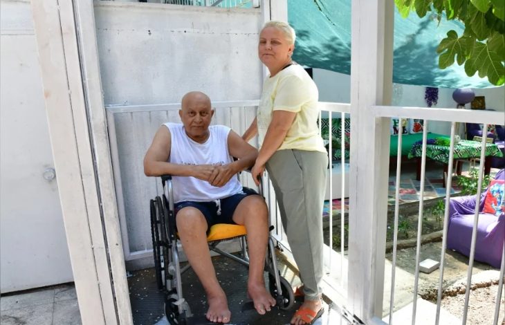 Görselde, açık hava ortamında tekerlekli sandalyede oturan yaşlı bir adam ve yanında ayakta duran bir kadın yer alıyor. Adam beyaz atlet ve koyu şort giymiş, çıplak ayaklı. Kadın ise açık renkli bir tişört ve pantolon giymekte, turuncu terlikleri var. İkili bir evin girişinde konumlanmış; etraflarında beyaz parmaklıklar bulunuyor. Arka planda yeşil bir tente ve renkli minderlerle dekore edilmiş bir bahçe alanı dikkat çekiyor.