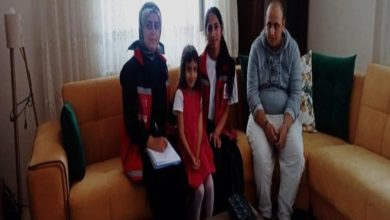 Görselde bir ev ortamında, sarı bir koltuk üzerinde oturan dört kişi görülüyor. Soldan sağa doğru iki kadın görevli kırmızı-siyah yelek giymiş; aralarında küçük bir kız çocuğu yer alıyor. En sağda ise gri kapüşonlu sweatshirt giymiş bir erkek oturmakta. Görevlilerden biri elinde bir defter tutuyor.