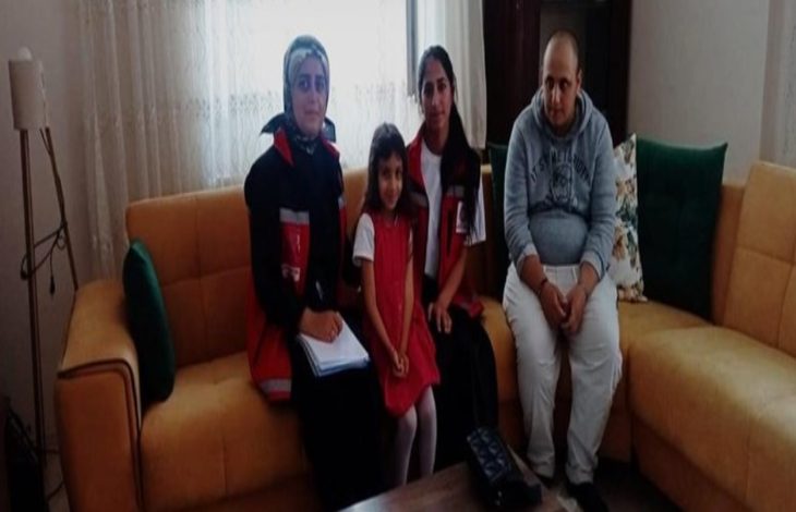 Görselde bir ev ortamında, sarı bir koltuk üzerinde oturan dört kişi görülüyor. Soldan sağa doğru iki kadın görevli kırmızı-siyah yelek giymiş; aralarında küçük bir kız çocuğu yer alıyor. En sağda ise gri kapüşonlu sweatshirt giymiş bir erkek oturmakta. Görevlilerden biri elinde bir defter tutuyor.