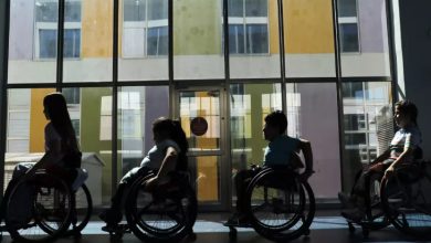 Görselde, tekerlekli sandalyedeki dört çocuk yan yana dizilmiş şekilde kapalı bir alanda görülüyor. Arkadan gelen ışık nedeniyle silüetleri öne çıkıyor. Büyük camlı kapının önünde, renkli duvarlarla çevrili bir binanın manzarası dikkat çekiyor.