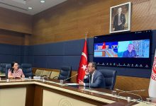 TBMM Engelli bireylerin Sorunlarının Araştırılması Komisyonu'nda salonunda, masada sağda Dr. Mehmet Muharrem Kasapoğlu solda ise Ayşe Sarı oturuyor. Masada mikrofon, su bardağı ve çeşitli belgeler yer almakta. Masanın arkasında, büyük bir ekran üzerinde bir Zoom toplantısı açık. Ekranda Mehmet Kızıltaş toplantıya dahil olmuş. Masanın sağ tarafında, Türkiye Büyük Millet Meclisi (TBMM) amblemini taşıyan bir bayrak bulunuyor.