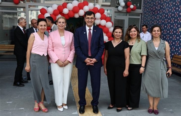 Görselde, Kütahya Belediyesi tarafından hayata geçirilen “Yamantürk Yaşlı ve Engelli Gündüz Yaşam Merkezi”nin açılış töreninden bir kare yer alıyor. Ön planda altı kişi – dört kadın, bir erkek ve bir kadın daha – resmi bir duruşla kameraya poz veriyor. Hepsi şık giyimli ve gülümseyerek açılışın coşkusunu yansıtıyor. Arkada kırmızı-beyaz balonlardan oluşan bir kemer, binanın girişini süslüyor.