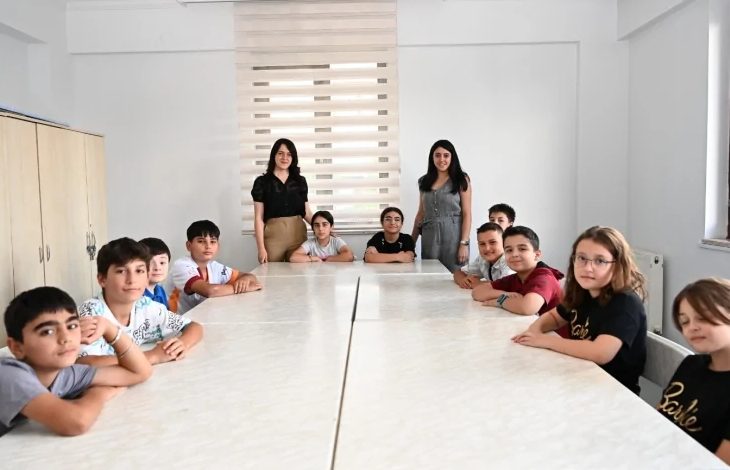 Görselde sınıf ortamında, dikdörtgen masaların etrafına oturmuş çocuklar yer alıyor. Masanın iki ucunda ise ayakta duran iki kadın bulunuyor. Çocuklar kameraya doğru bakıyor, sınıf düzeninde bir etkinlik havası var. Arka planda pencere ve kapalı stor perde görünüyor.