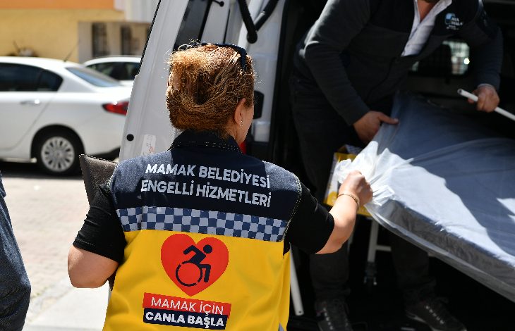 Görselde, sırtında “Mamak Belediyesi Engelli Hizmetleri” yazan bir yelek giymiş görevli, bir araca eşya (muhtemelen yatak veya medikal malzeme) yerleştirmeye yardım ediyor. Yeleğin üzerinde tekerlekli sandalye sembolü ve “Mamak için canla başla” ifadesi bulunuyor. Arka planda başka bir görevli ve park etmiş araçlar görünüyor.