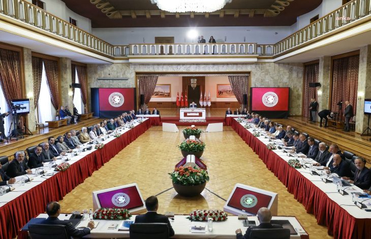 Türkiye Büyük Millet Meclisi büyük toplantı salonunda, TBMM Başkanı Numan Kurtuluş'un yönettiği toplantıda farklı siyasi partilerden gelen çok sayıda milletvekili ve bürokrat U şeklinde dizilmiş uzun masalarda katılımcı olarak bulunuyor.