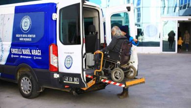 Görselde, engelli bireylerin taşınması için özel olarak tasarlanmış bir “Engelli Nakil Aracı” görülüyor. Aracın arka kapısı açık ve tekerlekli sandalyedeki bir kişi, lift sistemiyle dikkatli bir şekilde araca bindiriliyor. Sandalyeyi iten bir görevli ya da refakatçi de yardıma eşlik ediyor. Aracın üzerinde “Sağlık ve Sosyal Hizmetler Daire Başkanlığı” ibaresi yer almakta. Arka planda ise cam cepheli büyük bir kamu binası görülüyor.