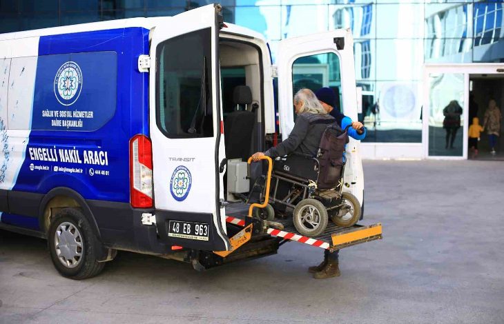 Görselde, engelli bireylerin taşınması için özel olarak tasarlanmış bir “Engelli Nakil Aracı” görülüyor. Aracın arka kapısı açık ve tekerlekli sandalyedeki bir kişi, lift sistemiyle dikkatli bir şekilde araca bindiriliyor. Sandalyeyi iten bir görevli ya da refakatçi de yardıma eşlik ediyor. Aracın üzerinde “Sağlık ve Sosyal Hizmetler Daire Başkanlığı” ibaresi yer almakta. Arka planda ise cam cepheli büyük bir kamu binası görülüyor.