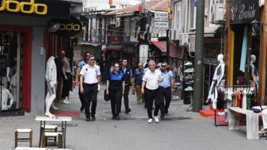 Görselde, bir grup zabıta görevlisi dar bir çarşı sokağında yürüyor. Ekip, üniformalarıyla birlikte sokakta denetim yapar halde görüntülenmiş. Her iki yanda butik ve giyim mağazaları bulunuyor; vitrinlerde giysi sergileyen mankenler dikkat çekiyor. Üst kısımda ise renkli sarkıt lambalar çarşıya canlı bir hava katıyor.