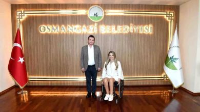 Görselde, bir erkek ve bir kadın resmi bir ortamda poz veriyor. Erkek takım elbise, kadın ise beyaz kıyafet giymiş ve tekerlekli sandalye kullanıyor. Arka planda “Osmangazi Belediyesi” yazısı, belediye logosu, Türk bayrağı ve belediye bayrağı yer alıyor. Zemin parlak ahşap kaplama.