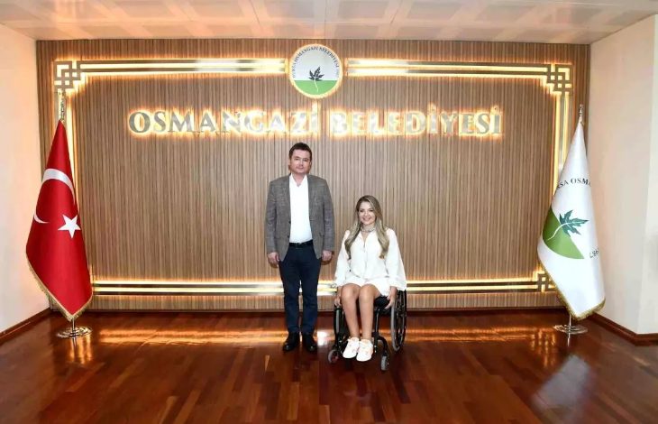 Görselde, bir erkek ve bir kadın resmi bir ortamda poz veriyor. Erkek takım elbise, kadın ise beyaz kıyafet giymiş ve tekerlekli sandalye kullanıyor. Arka planda “Osmangazi Belediyesi” yazısı, belediye logosu, Türk bayrağı ve belediye bayrağı yer alıyor. Zemin parlak ahşap kaplama.