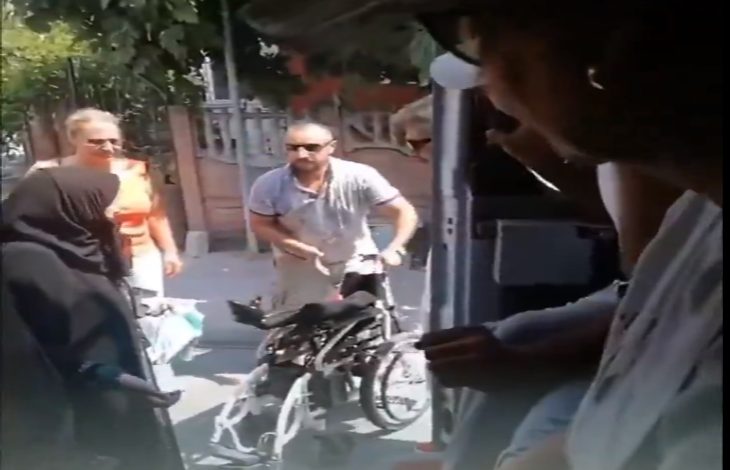 Görselde, dışarıda duran bir adam katlanmış bir tekerlekli sandalyeyi tutarken, çevresinde birkaç kişi bulunuyor. Fotoğraf, bir aracın içinden dışarıya doğru çekilmiş ve sokak ortamı net şekilde görülüyor.