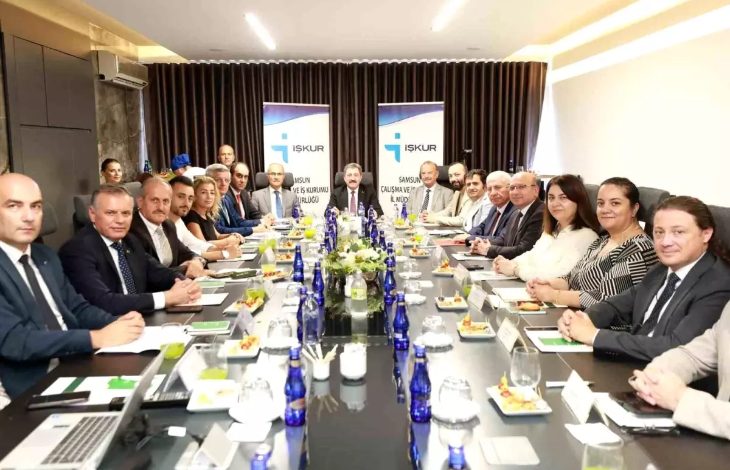 Görselde, uzun bir toplantı masası etrafında toplanmış kalabalık bir grup görülüyor. Katılımcılar resmi kıyafetler içinde ve çoğu ciddi bir ifadeyle kameraya bakıyor. Masada su şişeleri, meyve tabakları, not defterleri ve isimlikler yer alıyor. Arka planda “İŞKUR” logolu iki pano ile “Samsun Çalışma ve İş Kurumu İl Müdürlüğü” yazıları dikkat çekiyor.