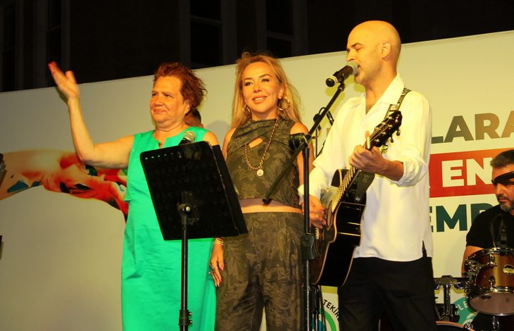 Görselde, sahnede üç kişi mikrofon önünde yer alıyor. Bir kadın yeşil elbiseyle el sallarken, ortadaki kadın gülümseyerek izleyiciye bakıyor. Yanlarında Murat Evgin ise gitar çalıyor ve şarkı söylüyor. Arka planda davulcu ve etkinlik afişinin bir kısmı görünüyor.