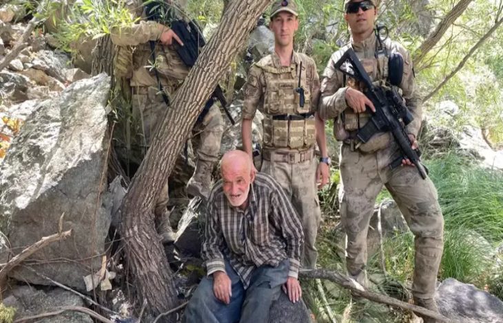 Görselde, kırsal bir bölgede kayalık ve ağaçlık alanda bulunan yaşlı bir adam yer alıyor. Üzerinde kareli gömlek ve koyu renk pantolon var, oldukça bitkin görünüyor. Yanında ve arkasında ise silahlı ve üniformalı jandarma personeli bulunuyor.