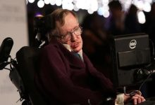 Görselde, ünlü teorik fizikçi Stephen Hawking yer alıyor. Elektrikli tekerlekli sandalyesinde oturmakta ve yanında, iletişim kurmak için kullandığı Intel destekli özel bilgisayar ekranı bulunuyor. Üzerinde koyu renk bir giysi var ve yüz ifadesi ciddi. Arka planda flu şekilde ışıklar ve insanlar görünüyor, bu da etkinlik tarzı bir ortam izlenimi veriyor.