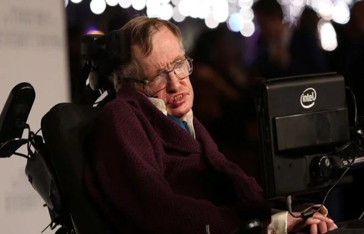Görselde, ünlü teorik fizikçi Stephen Hawking yer alıyor. Elektrikli tekerlekli sandalyesinde oturmakta ve yanında, iletişim kurmak için kullandığı Intel destekli özel bilgisayar ekranı bulunuyor. Üzerinde koyu renk bir giysi var ve yüz ifadesi ciddi. Arka planda flu şekilde ışıklar ve insanlar görünüyor, bu da etkinlik tarzı bir ortam izlenimi veriyor.