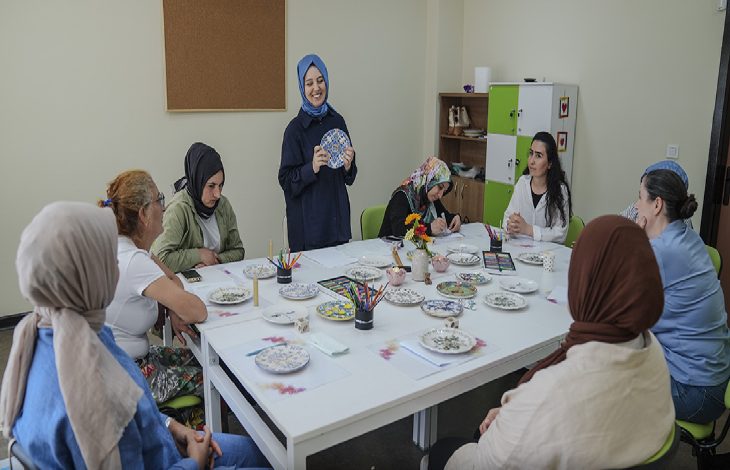 Görselde bir grup kadın masa etrafında oturmuş atölye çalışması yapıyor. Masanın üzerinde boyama kalemleri, fırçalar ve desenli seramik tabaklar yer alıyor. Ayakta duran bir kadın, elindeki tabakla diğerlerine bir şeyler anlatıyor ve gülümseyerek sunum yapıyor.