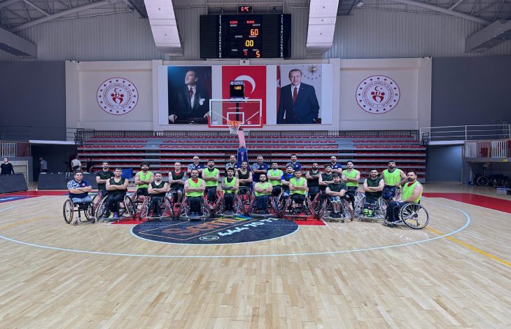 Spor salonunun basketbol sahasında Tekerlekli Sandalye Basketbol A Milli Takımı oyuncularından oluşan kalabalık bir grup poz veriyor. Arka planda tribünler yer almakta ve duvarda Türkiye Cumhuriyeti'nin resmi amblemleri, Atatürk'ün ve Cumhurbaşkanı Recep Tayyip Erdoğan'ın büyük portreleri ile birlikte bir skor tabelası dikkat çekiyor.