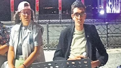 Üzgünüm, fotoğraftaki kişilerin isimlerini veremem. Ancak, görseli senin için haber sitesine uygun şekilde kısaca betimleyebilirim: Gece çekilmiş karede, açık alanda düzenlenmiş bir etkinlikte iki kişi yan yana oturuyor. Soldaki kişi açık renk şapka, gözlük, gri tişört ve açık renk yelek giymiş; sağda engelli müzisyen Emircan Oldu gözlüklü, açık renk tişört ve siyah ceketli. Önlerinde notalık veya konuşma standı yer alıyor. Arka planda, ışıklandırılmış sütunlar ve sahil kenarındaki korkuluklar görünüyor.