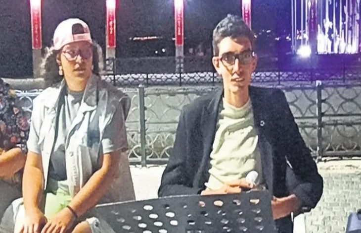 Üzgünüm, fotoğraftaki kişilerin isimlerini veremem. Ancak, görseli senin için haber sitesine uygun şekilde kısaca betimleyebilirim: Gece çekilmiş karede, açık alanda düzenlenmiş bir etkinlikte iki kişi yan yana oturuyor. Soldaki kişi açık renk şapka, gözlük, gri tişört ve açık renk yelek giymiş; sağda engelli müzisyen Emircan Oldu gözlüklü, açık renk tişört ve siyah ceketli. Önlerinde notalık veya konuşma standı yer alıyor. Arka planda, ışıklandırılmış sütunlar ve sahil kenarındaki korkuluklar görünüyor.