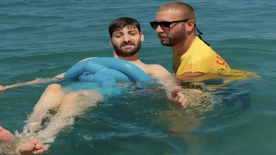 Soldaki genç erkek, denizde sırtüstü yatar pozisyonda ve rahat bir şekilde suyun üzerinde duruyor. Üzerinde mavi renkte havuz makarnası bulunuyor. Sağdaki sarı tişörtü ve siyah güneş gözlüklü yardımcı görevli erkek, engelli kişiyi su üstünde tutuyor.