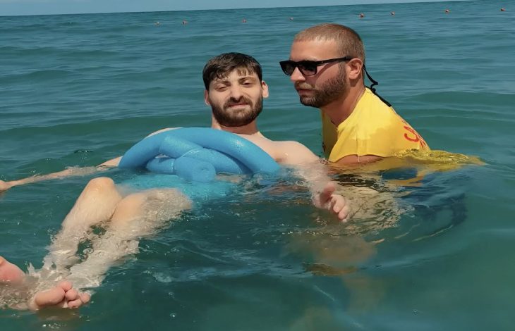 Soldaki genç erkek, denizde sırtüstü yatar pozisyonda ve rahat bir şekilde suyun üzerinde duruyor. Üzerinde mavi renkte havuz makarnası bulunuyor. Sağdaki sarı tişörtü ve siyah güneş gözlüklü yardımcı görevli erkek, engelli kişiyi su üstünde tutuyor.