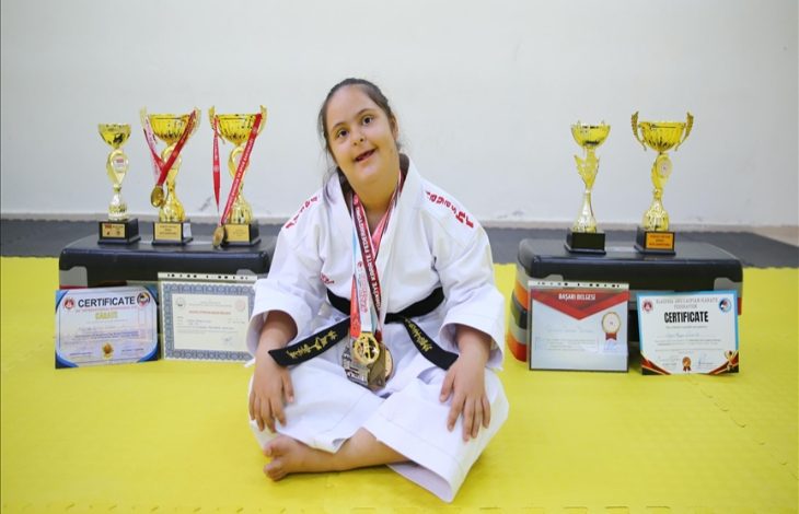 Görselde, karate kıyafeti giymiş genç bir kız yerde oturuyor. Boynunda kazandığı madalyalar asılı. Arka planda ise çok sayıda kupa ve başarı sertifikası sıralanmış. Kızın gülümseyen ifadesi, spor yoluyla elde ettiği başarı ve gururu yansıtıyor.