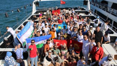 Geniş bir teknenin üst güvertesinde, farklı yaş ve milletlerden oluşan özel sporcular ile kafileleri bir araya gelmiş. Katılımcılar kameraya doğru neşeyle el sallıyor, poz veriyorlar. Kalabalığın ortasında ve önünde çeşitli bayraklar dikkat çekiyor: Türkiye, Özbekistan, Kazakistan ve Kırgızistan bayrakları gururla tutuluyor. Üst güvertenin arka kısmında Türk bayrağı dalgalanıyor.