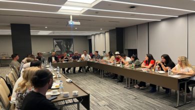 Modern bir toplantı salonunda, dikdörtgen masalar U şeklinde dizilmişken, farklı yaş ve cinsiyetlerden oluşan yaklaşık 20 kişilik federasyon yetkilileri toplantı yapıyor. Masaların üzerinde su şişeleri, kahve fincanları, atıştırmalıklar ve not defterleri yer alıyor.