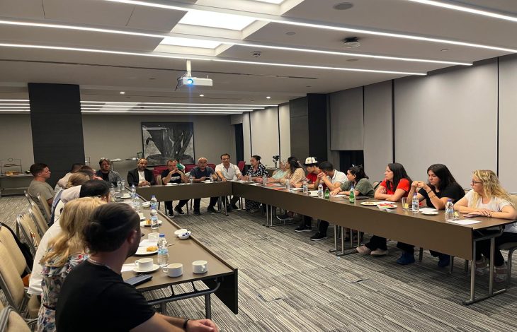 Modern bir toplantı salonunda, dikdörtgen masalar U şeklinde dizilmişken, farklı yaş ve cinsiyetlerden oluşan yaklaşık 20 kişilik federasyon yetkilileri toplantı yapıyor. Masaların üzerinde su şişeleri, kahve fincanları, atıştırmalıklar ve not defterleri yer alıyor.