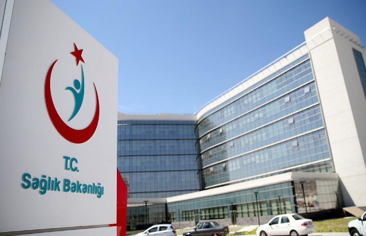 Görselde, modern mimariye sahip büyük bir kamu binası ve önünde yer alan T.C. Sağlık Bakanlığı tabelası görülüyor. Tabelada kırmızı hilal ve yıldız sembolüyle birlikte stilize bir insan figürü bulunuyor. Binanın cephesi tamamen camla kaplı ve oldukça çağdaş bir yapıyı yansıtıyor. Ön kısımda birkaç otomobil park halinde. Hava açık ve güneşli.