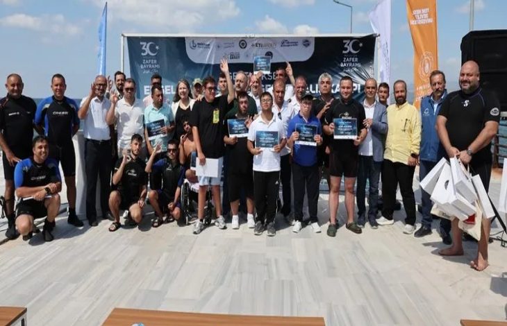 Görselde açık havada bir grup insan toplu fotoğraf çekiliyor. Ön sırada ellerinde sertifika tutan engelli sporcular ve yanlarında antrenörler bulunuyor. Arka planda etkinlik afişi ve bayraklar yer alıyor. Katılımcıların çoğu spor kıyafetli, bazılarının dalış kıyafeti giydiği görülüyor.