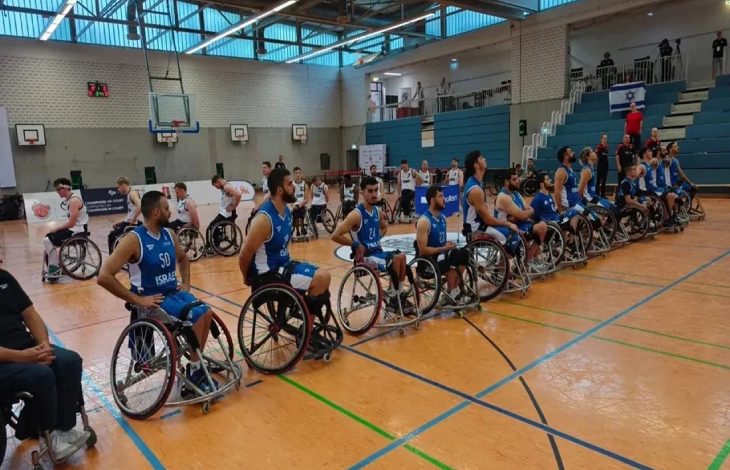 Mavi forma ile sahada olan İsrail Milli Takımına arkalarında bulunan beyaz formalı İngiltere Tekerlekli Sandalye Basketbol Milli Takımı sporcuları sırtlarını dönerken.