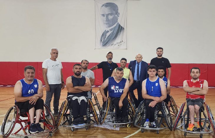 Görselde, bir spor salonunda tekerlekli sandalyedeki basketbol oyuncuları ve ayakta duran kişiler toplu halde poz veriyor. Oyuncuların üzerinde mavi ve kırmızı formalar var. Arka planda Atatürk portresi asılı. Grup, yeni sezon öncesi takım pozu verir gibi kameraya bakıyor.