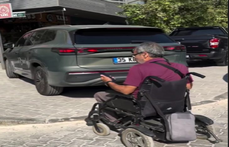 Görselde, elektrikli tekerlekli sandalye kullanan bir birey kaldırıma çıkmak isterken, kaldırımın girişini kapatan bir aracın (TOGG) yüzünden ilerleyemiyor. Kişi, elinde telefonuyla bu duruma tepkisini gösteriyor gibi görünüyor. Araç, engelli bireyin geçiş hakkını engelleyerek ciddi bir erişilebilirlik sorununa yol açıyor.