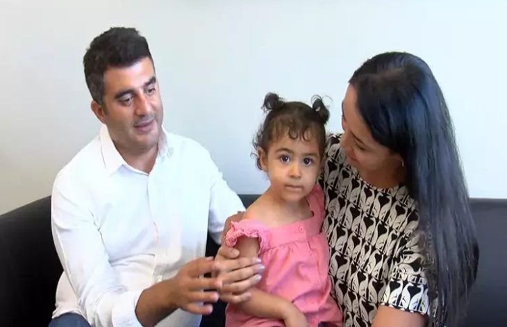 Fotoğrafta bir aile yer alıyor: solda beyaz gömlek giymiş bir baba, ortada pembe elbise giymiş küçük bir kız çocuğu ve sağda siyah-beyaz desenli bluz giymiş bir anne bulunuyor. Baba, kızının omzuna dokunurken, anne şefkatle çocuğuna bakıyor. Kız çocuğu ise doğrudan kameraya bakıyor.