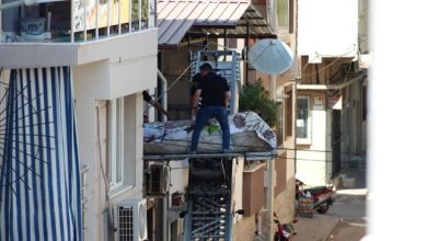 Görselde, dar bir sokakta bir apartman dairesinin penceresinden dışarı doğru uzanan eşya taşıma asansörü üzerinde, bir kişi yatay şekilde sedyeye benzer bir düzlemde taşınıyor. Üzerinde çarşaf örtülü bedenin cenaze olduğu anlaşılıyor. Yanında bir görevli ya da taşıyıcı bulunmakta. Alt sokakta motorlar ve evler dikkat çekiyor.
