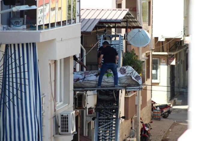 Görselde, dar bir sokakta bir apartman dairesinin penceresinden dışarı doğru uzanan eşya taşıma asansörü üzerinde, bir kişi yatay şekilde sedyeye benzer bir düzlemde taşınıyor. Üzerinde çarşaf örtülü bedenin cenaze olduğu anlaşılıyor. Yanında bir görevli ya da taşıyıcı bulunmakta. Alt sokakta motorlar ve evler dikkat çekiyor.